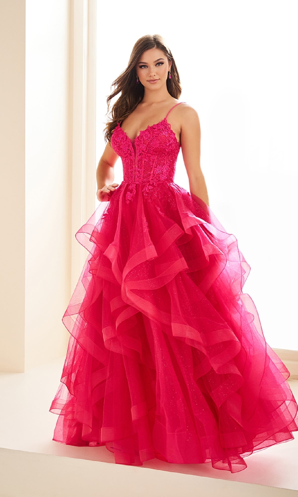 A woman stands indoors wearing the Ellie Wilde EW36203 Glitter Tulle Prom Ball Gown, featuring bright pink ruffled layers, thin straps, a fitted embroidered corset bodice, and an elegant adjustable lace-up back.